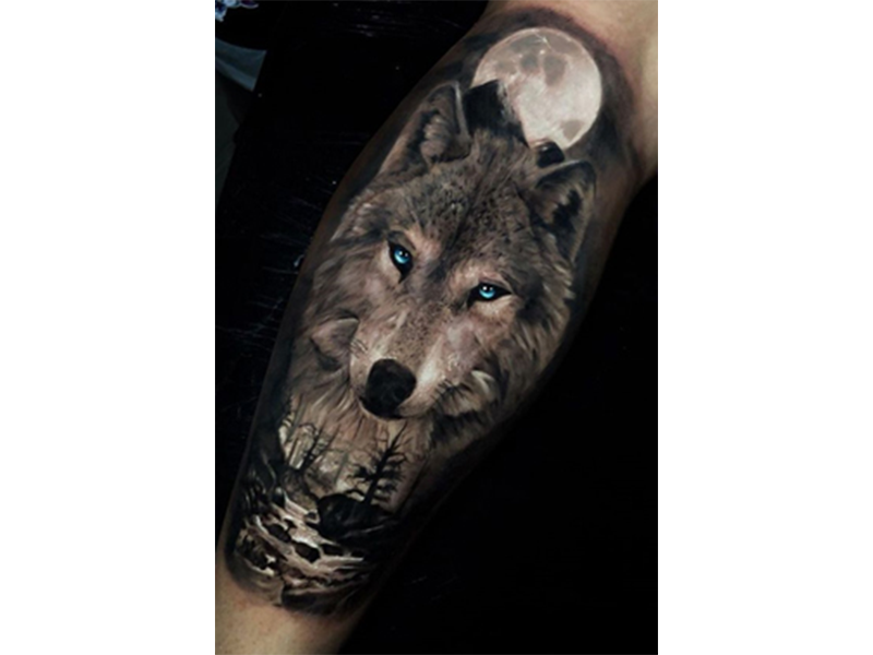 Black and white calf tattoo of a wolf with blue eyes in front of a full moon and above a rock-filled stream with nearby trees